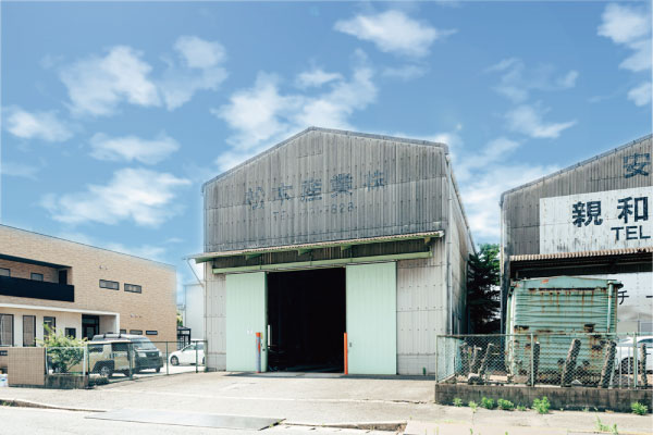 雲が散らばる青空の下、別の倉庫とベージュの建物の間に、入り口の開いた金属製の倉庫が建っている。近くに駐車中の車と緑が見える。.