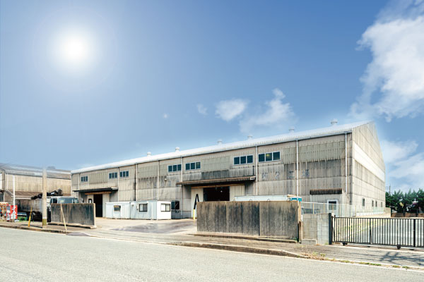 晴れ渡った青空の下、波型の金属外壁を持つ大きな長方形の工業用倉庫ビルが太陽とともに写っている。倉庫には複数の大きな搬入口があり、舗装された道路の脇に位置している。.