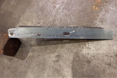 長方形の金属棒に3つの穴が開けられ、すり減った灰色の工業用表面に置かれている。棒の端は面取りされ、表面にはかすかにマークが見える。.