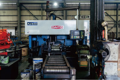 DAITO DS400産業用機械が、金属部品やコンテナ、設備に囲まれた工場内に立っている。コントロールパネルが見え、整理整頓され使用されているように見える。.