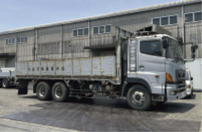 風化した荷台を持つ銀色の平台トラックが、いくつもの窓と波形の外壁を持つ大きな工業用倉庫の前に駐車している。.