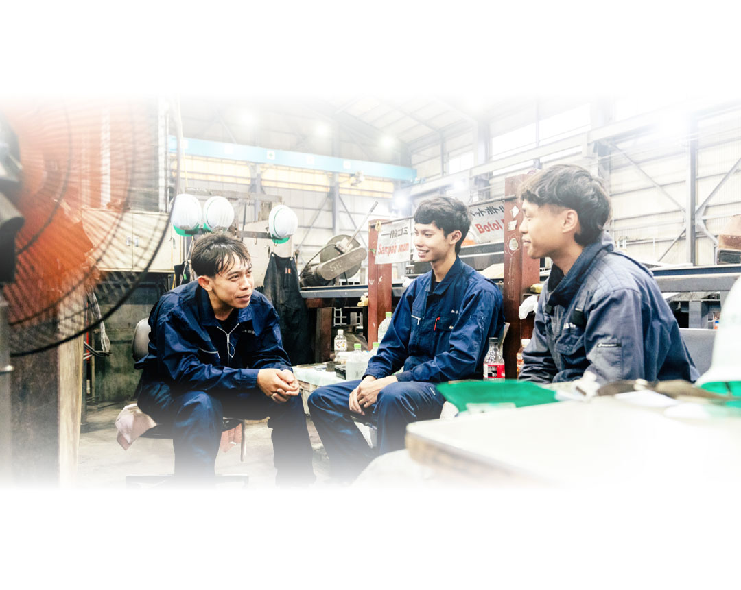 青い作業服を着た3人の男性が、工業用作業場の中で座って話している。背景にはヘルメットと工具が見え、照明は明るく、縁には少しぼやけた白いケラレがある。.