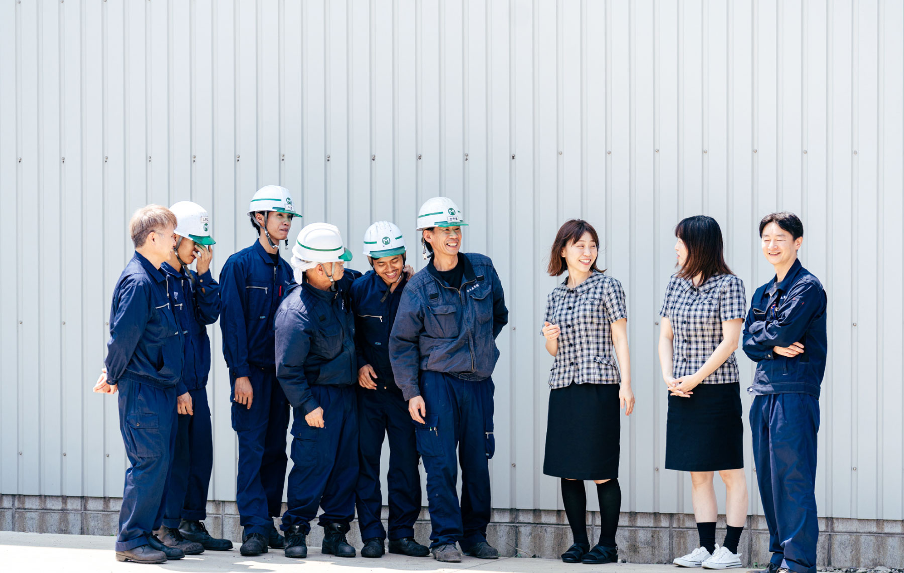 青い作業服に白いヘルメット姿の6人と、チェックのシャツに黒いスカート姿の2人、計8人のグループが波板の壁の前に立って話している。何気ない会話をしているようだ。.