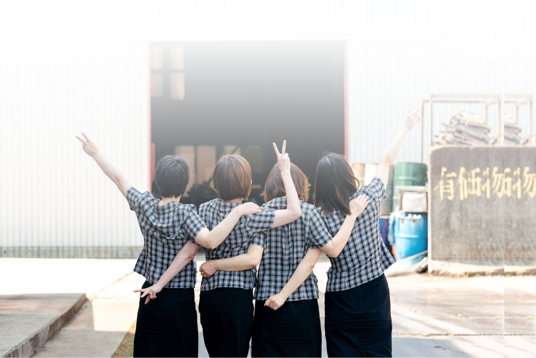 4人がカメラに背を向け、互いの肩に腕を回して並んでいる。彼らは同じようなチェックのシャツを着てピースサインをしており、工業的な特徴を持つ建物に面している。.