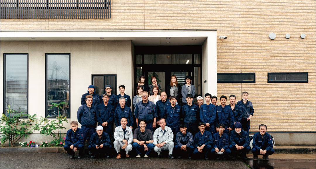 大きな窓とドアのあるベージュのレンガ造りの建物の前で、紺色の作業服を着た男性を中心とした31人のグループが記念撮影に応じる。後列にはカジュアルやビジネスウェアに身を包んだ女性も立っている。.