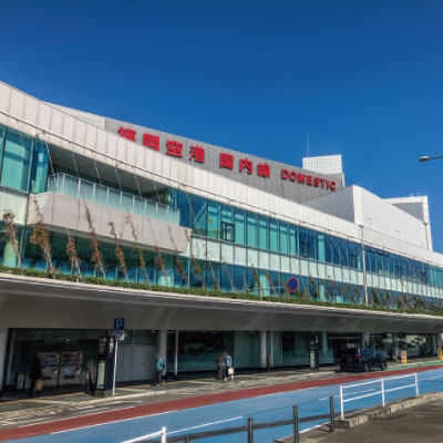 2階建ての空港ターミナルビル。大きなガラス窓があり、上空には青空が広がり、赤い看板には日本語と英語で「DOMESTIC」と書かれている。入り口付近には車や人が見える。.