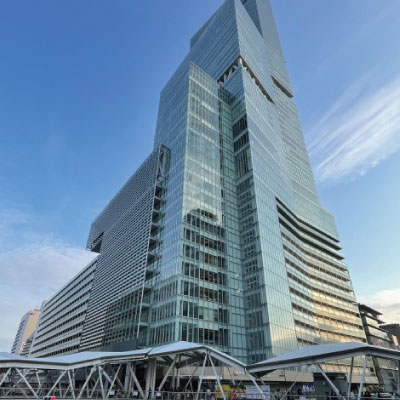 澄み切った青空を背景に、角ばった千鳥格子のモダンなガラス張りの高層ビルが聳えている。タワーの麓には屋根付きの歩道と低い建物がいくつか見える。.