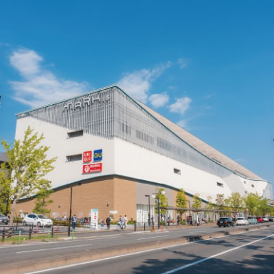 晴れた日に見える、白と茶色の外壁を持つ近代的な大型ショッピングセンター。通り沿いには車が停まり、歩道を人々が歩き、頭上には雲が散らばる青空が広がっている。.