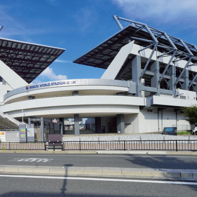 晴れた日の静かな通りに、IMAZU WORLD STADIUMと書かれた角ばった屋根の近代的なスポーツスタジアムが建っている。白い壁、大きな張り出し、いくつもの入り口。.