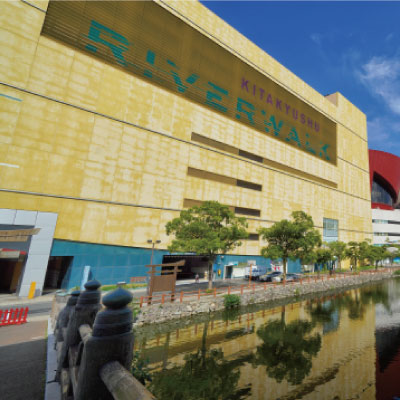 北九州リバーウォークと書かれた大きな黄色い建物が運河に映っている。水面には木々が立ち並び、ビルの入り口には歩道が通っている。手前には橋と手すりが見える。.