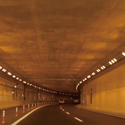 黄色い照明、車線表示、数台の先行車があるカーブした道路トンネル内からの眺め。トンネルの壁と天井が照らされ、小さな交通コーンが車線を区切っている。.