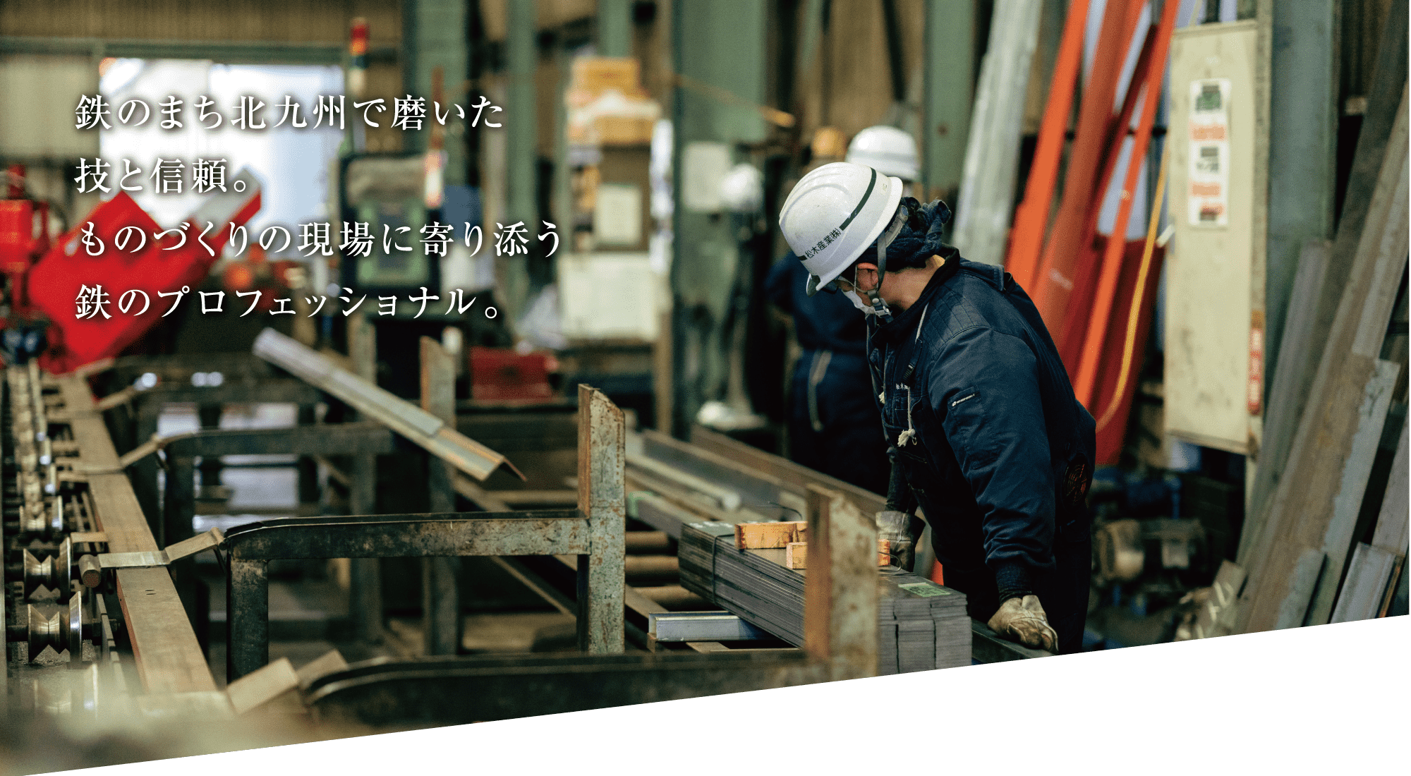 保護ヘルメットをかぶり、黒っぽい制服を着た作業員が、機械や工具を背景に、工業作業場で金属の梁を検査している。画像の左側に日本語の文字が重なっている。.