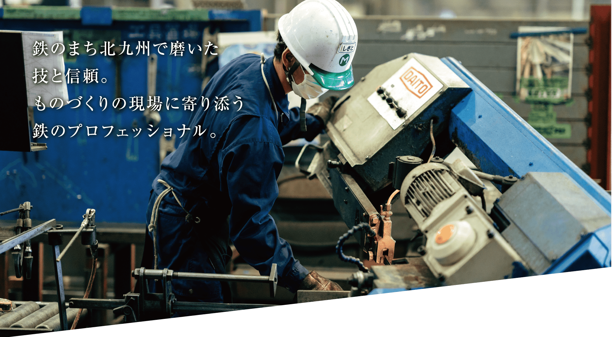 青いつなぎ服に白いヘルメット姿の作業員が工場で金属加工機を操作している。画像の左側に日本語の文字が重なっている。.