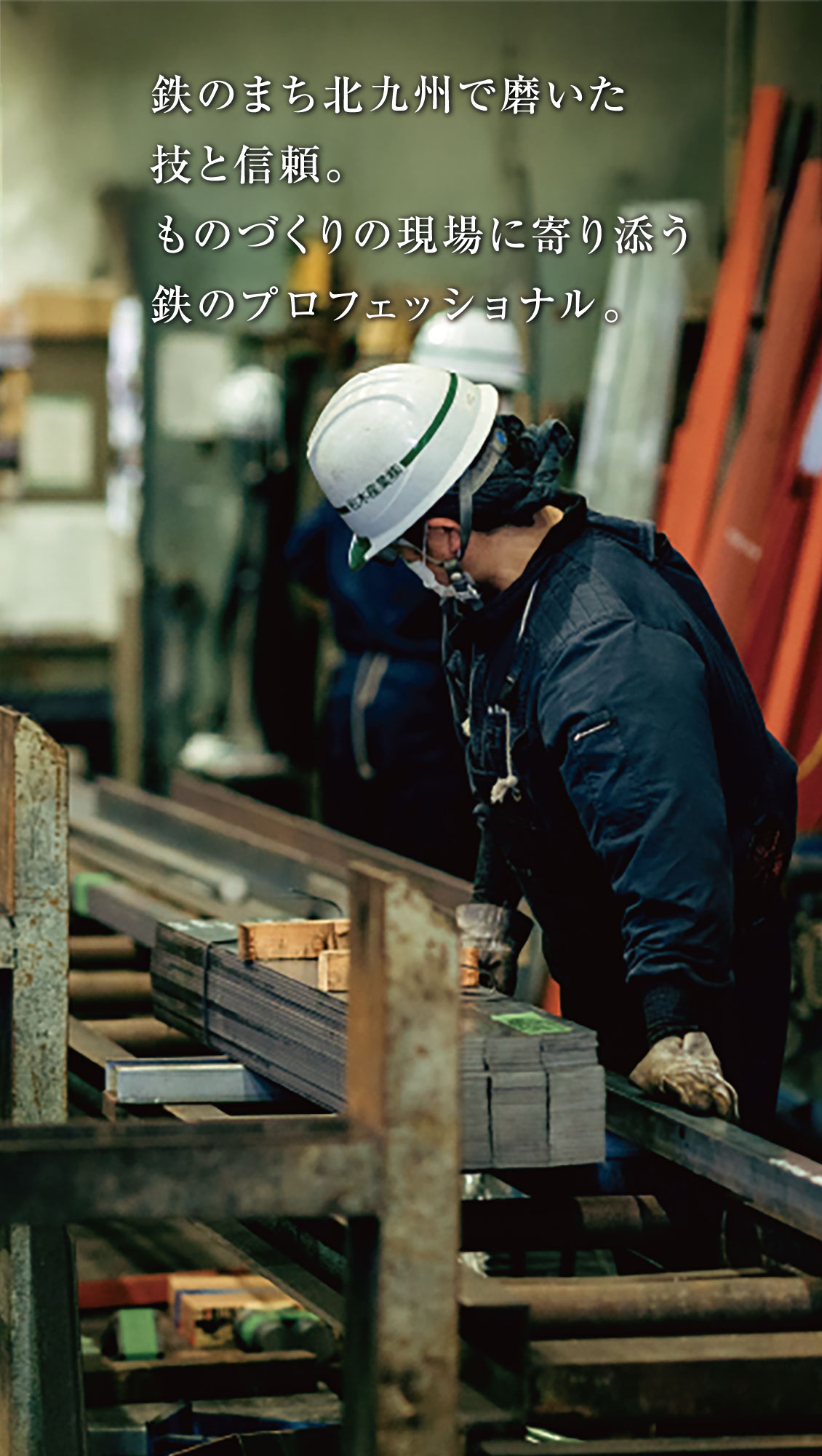 白い安全ヘルメットをかぶり、黒っぽい作業着と手袋をはめた人が、金属工場で金属棒を扱いながら作業している。上部に日本語の文字が重なっている。.